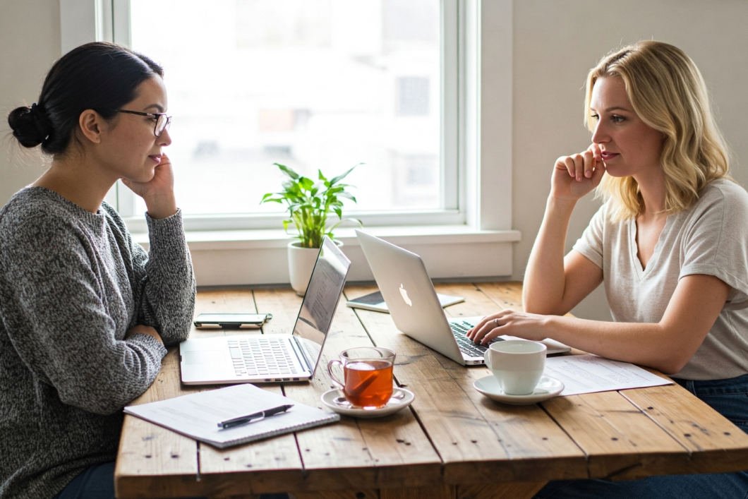 Two women collaborating on laptops at a wooden table with tea and papers, representing smart event planning and budgeting discussions.